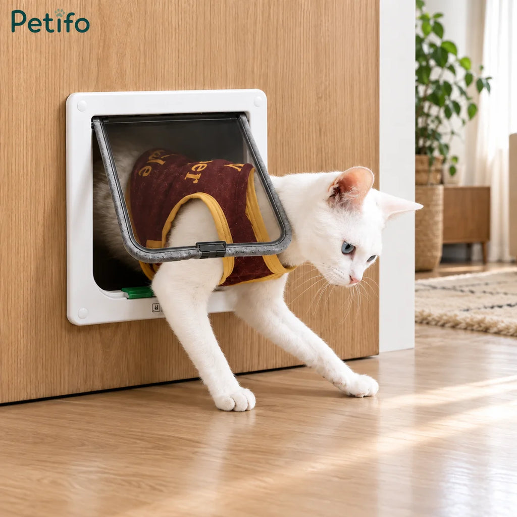 White 4-way locking cat flap installed on a wooden door with a white cat wearing a maroon and yellow vest stepping through.