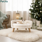 A cute cat resting on a plush, round elevated pet bed positioned next to a beautifully decorated Christmas tre