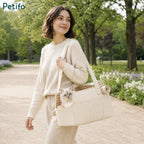 A woman in a cream-colored sweater walking outdoors on a path, carrying a beige Petifo pet carrier bag containing a fluffy cat.