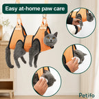 A gray cat suspended in an orange Petifo grooming hammock while someone clips its nails and cleans its face and ears.
