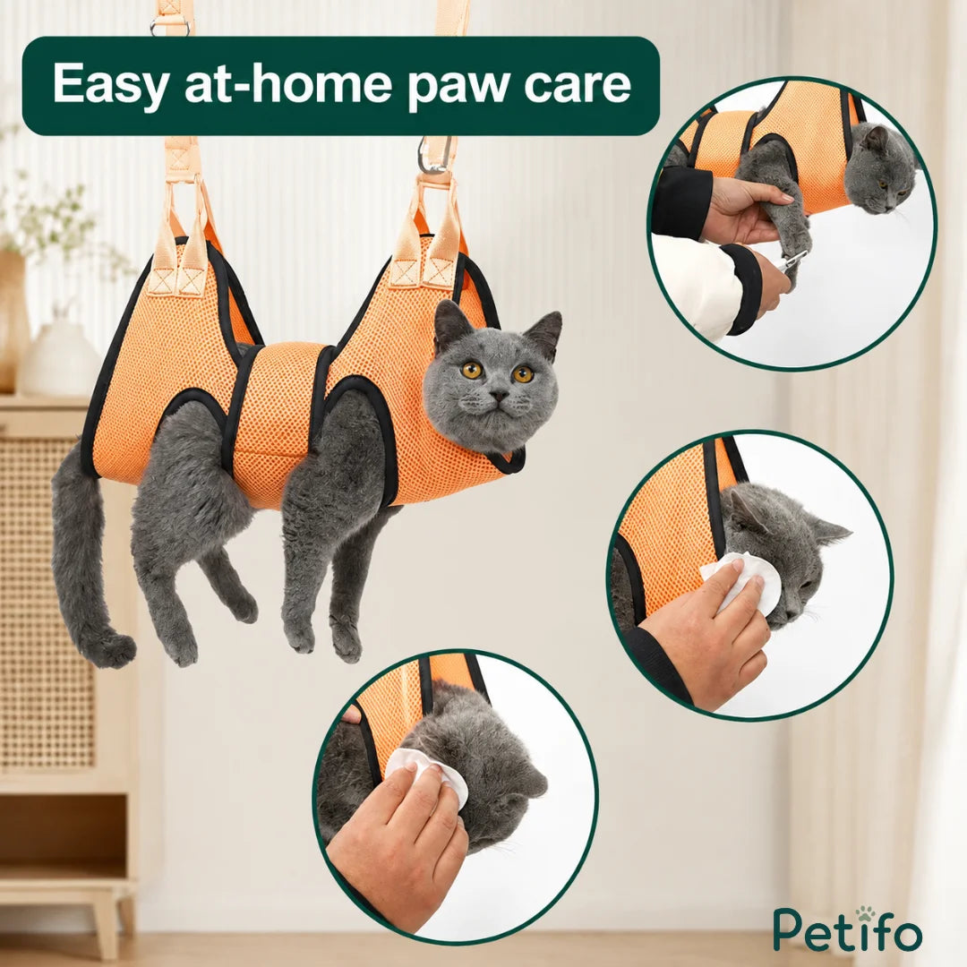 A gray cat suspended in an orange Petifo grooming hammock while someone clips its nails and cleans its face and ears.