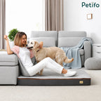 A happy woman sitting on a comfortable, large gray and white pet bed indoors, resting with her golden retriever.