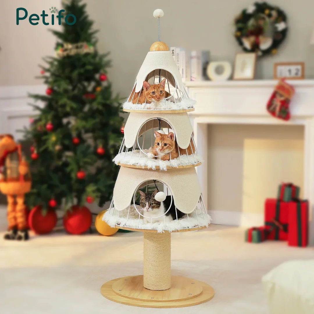 Three cats relaxing on different levels of a cream-colored, Christmas tree-themed cat tower placed in a cozy, decorated living room.