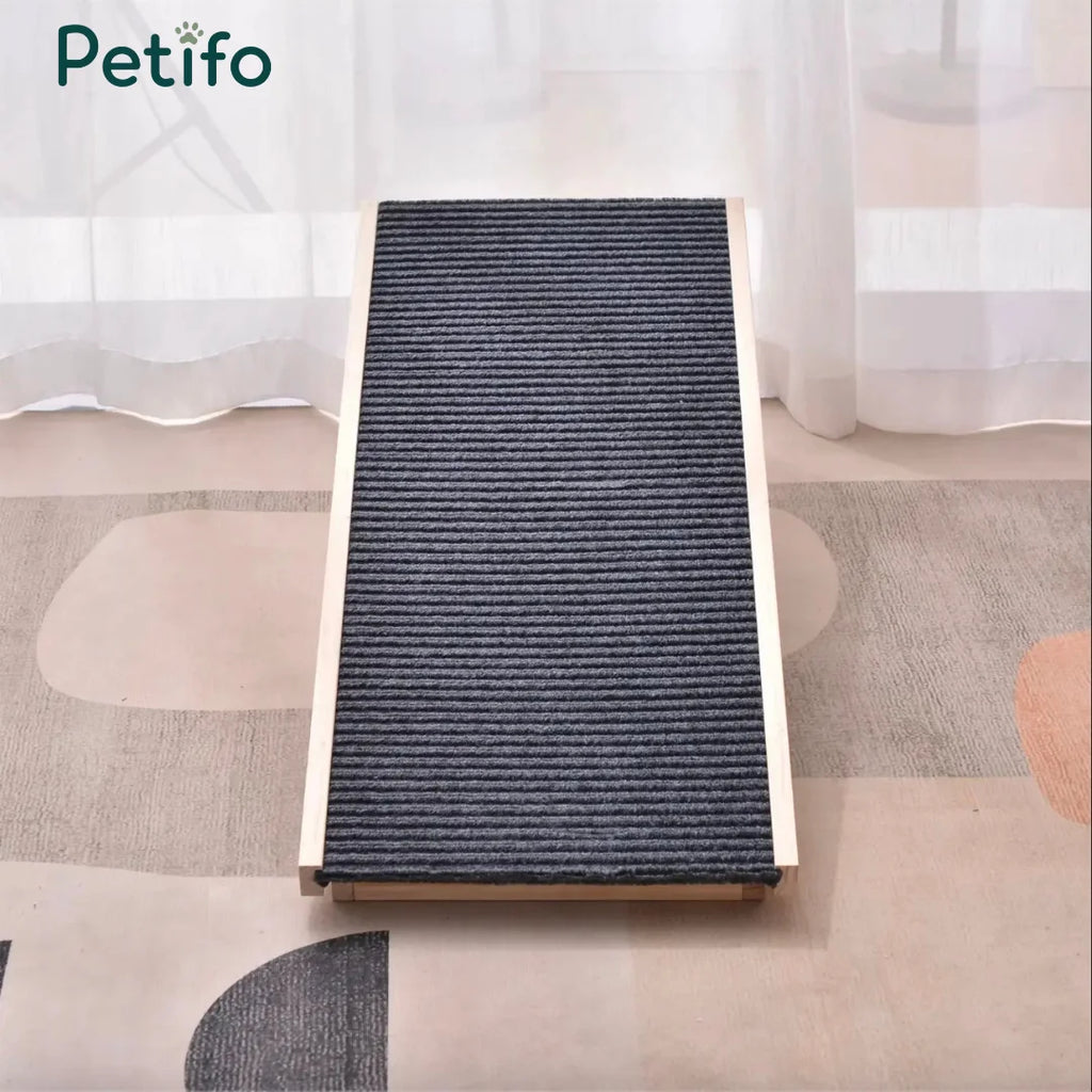 A wooden pet ramp with a dark gray, ribbed non-slip surface positioned on a patterned rug in a bright room. 
