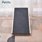 A wooden pet ramp with a dark gray, ribbed non-slip surface positioned on a patterned rug in a bright room. 
