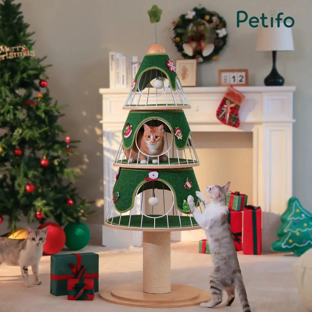 Three cats interacting with a Christmas tree-shaped, three-tier cat tower decorated with ornaments in a festive living room setting.