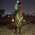 Equestrian rider on a brown horse equipped with a green LED breastplate at night.