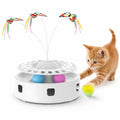 An energetic orange kitten playing with a white 3-in-1 interactive cat toy featuring spinning butterflies and colorful rolling balls.