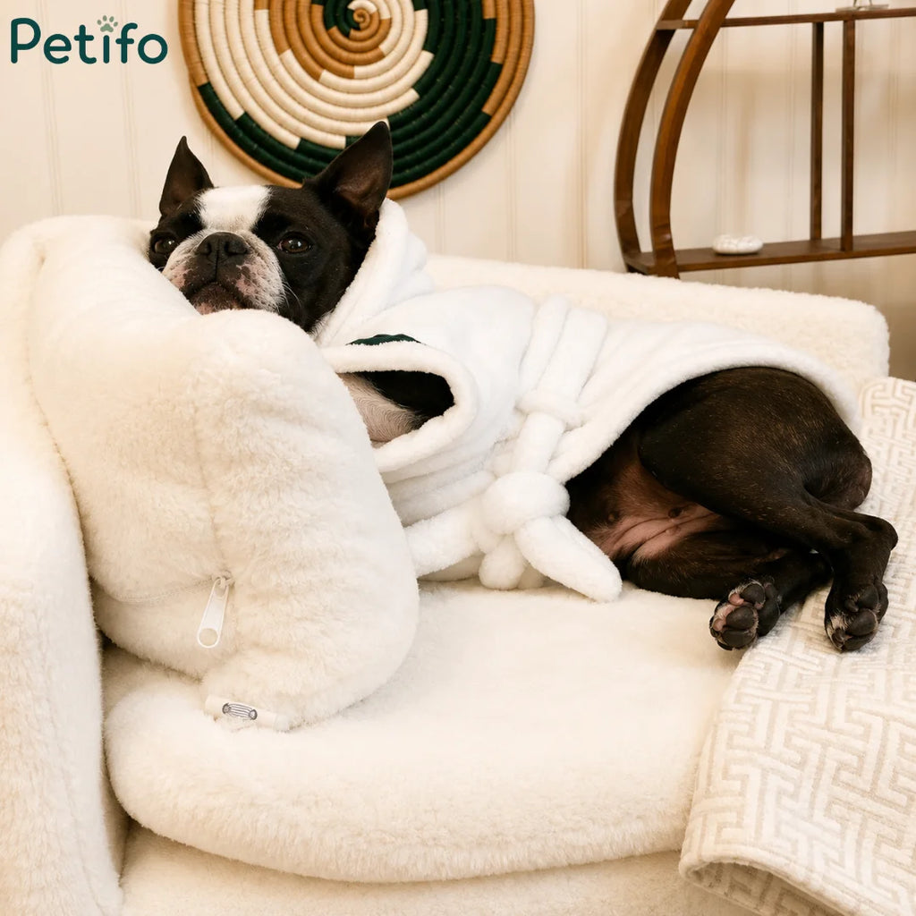 A Boston Terrier in a bathrobe relaxing on a luxury white plush pet sofa in a decorated living room.