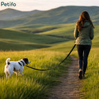 Woman walking a Jack Russell terrier on a black long training leash along a scenic green hill path at sunset.