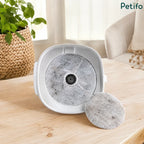 White pet water fountain base with a used filter installed and an extra replacement filter disc resting on a wooden table.